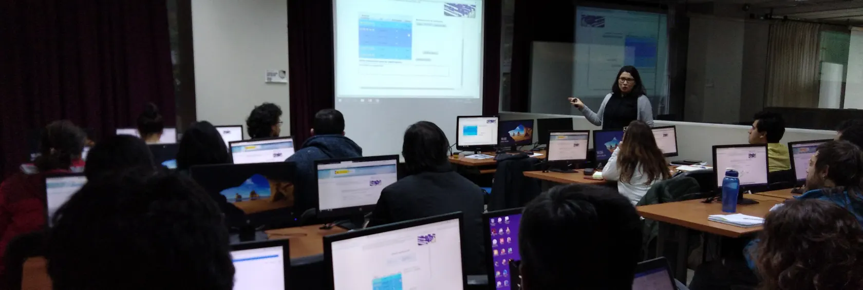 Profesora dando su clase y exponiendo ante los alumnos donde estan sentados cada uno frente a un computador